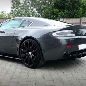 Gray sports car (Aston Martin) with large black multi-spoke wheels parked on a paved lot, rear three-quarter view.