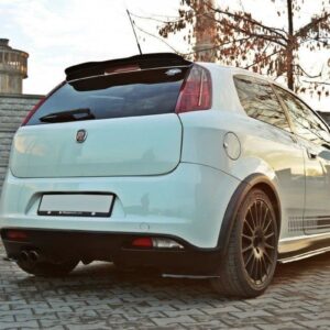 White hatchback car with custom dark alloy wheels, rear spoiler, and dual exhausts parked on a brick driveway. Moment captures a sporty exterior modification.