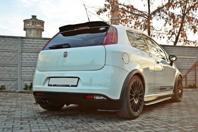 White hatchback car with custom dark alloy wheels, rear spoiler, and dual exhausts parked on a brick driveway. Moment captures a sporty exterior modification.