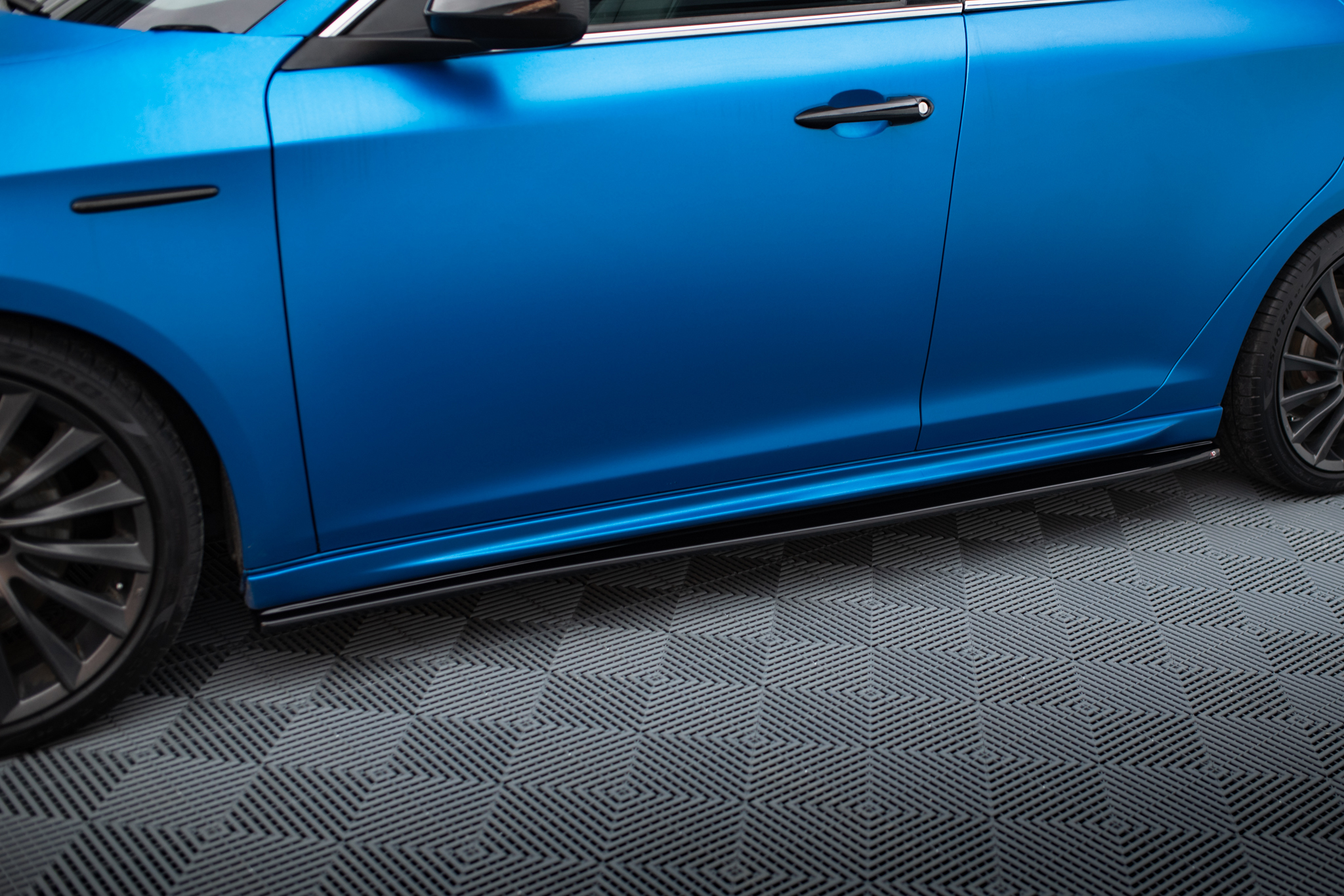 pol_po_Side-Skirts-Diffusers-V-1-suitable-for-Alfa-Romeo-Giulietta-601_2jpg - OPSHOLDERS Blue car door with a black handle, wheel visible to the left, on a patterned floor.