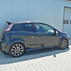 Black compact hatchback with a rear spoiler parked on a paved lot beside a light industrial building with a large garage door, sunlit scene.