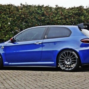 pol_po_Side-Skirts-Diffusers-suitable-for-Alfa-Romeo-147-GTA-76_3jpg - OPSHOLDERS Blue sporty hatchback with large aftermarket wheels and a rear spoiler, parked on a brick driveway with a hedge in the background.