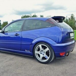Blue compact sport hatchback with large silver alloy wheels parked on a paved surface in an open area with trees in the background, rear spoiler visible.