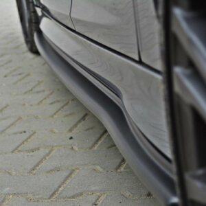 Close-up of a dark car's lower side panel with a noticeable dent along the rocker, resting on a paved brick surface.