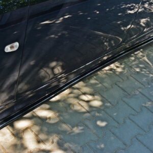 Black car side with a low side skirt and large alloy wheel, parked on a paved surface with dappled shadows