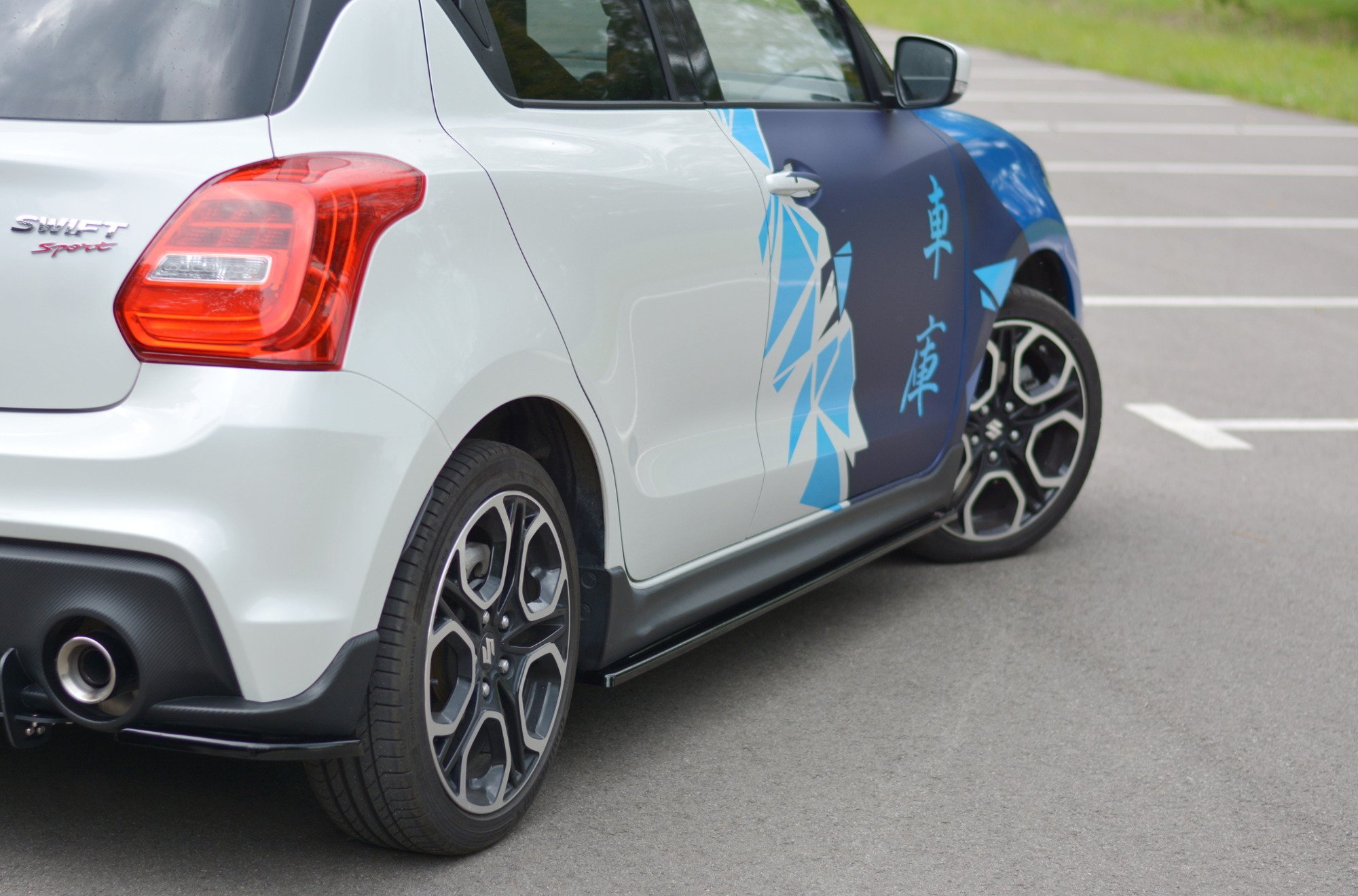 pol_po_Side-Skirts-Diffusers-suitable-for-Suzuki-Swift-Sport-Mk6-8264_3jpg - OPSHOLDERS White hatchback with blue geometric wrap along the side, showing rear taillight and alloy wheels in a parking lot.