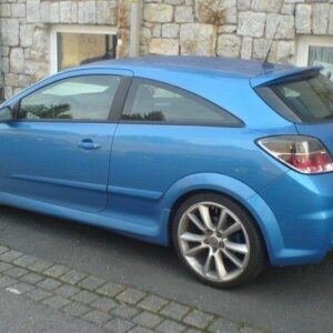 Blue two-door hatchback parked on a street with a stone wall behind, profile view on the left side of the image.