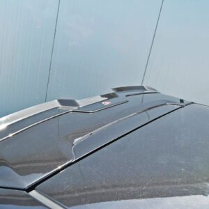 Close-up of a dark car roof showing a rear spoiler and roof-mounted antenna against a blue wall outside.