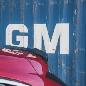 Close-up of a pink car's rear with a blue, weathered metal wall and large white letters in the background.