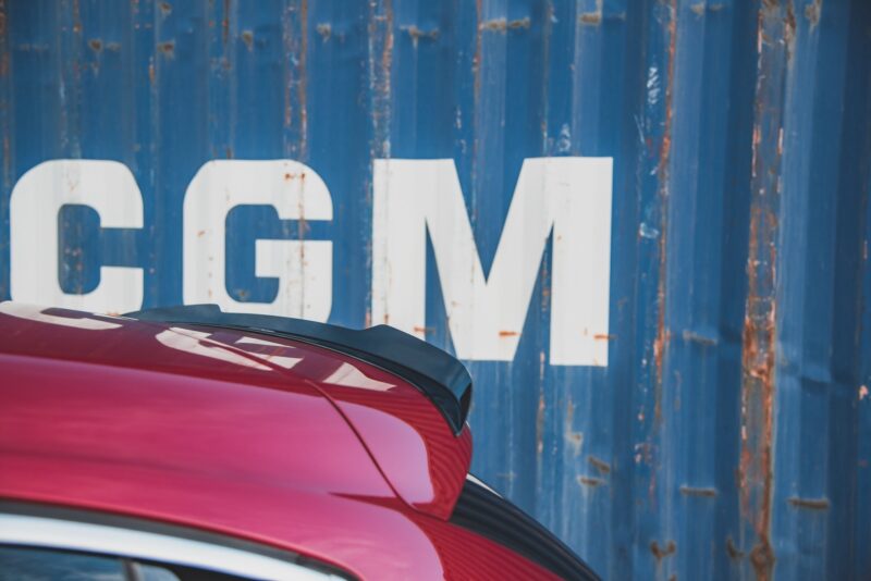 Close-up of a pink car's rear with a blue, weathered metal wall and large white letters in the background.