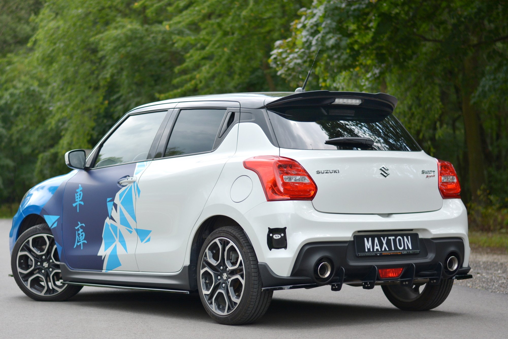 pol_po_Spoiler-Cap-suitable-for-Suzuki-SWIFT-6-SPORT-8255_2jpg - OPSHOLDERS Rear three-quarter view of a white Suzuki SUV with blue graphics, parked on a road with green trees in the background, Maxton license plate visible on the tailgate.
