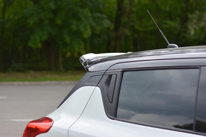pol_po_Spoiler-Cap-suitable-for-Suzuki-SWIFT-6-SPORT-8255_3jpg - OPSHOLDERS Rear left corner of a silver SUV, showing the roofline and antenna against a blurred green background in a parking lot